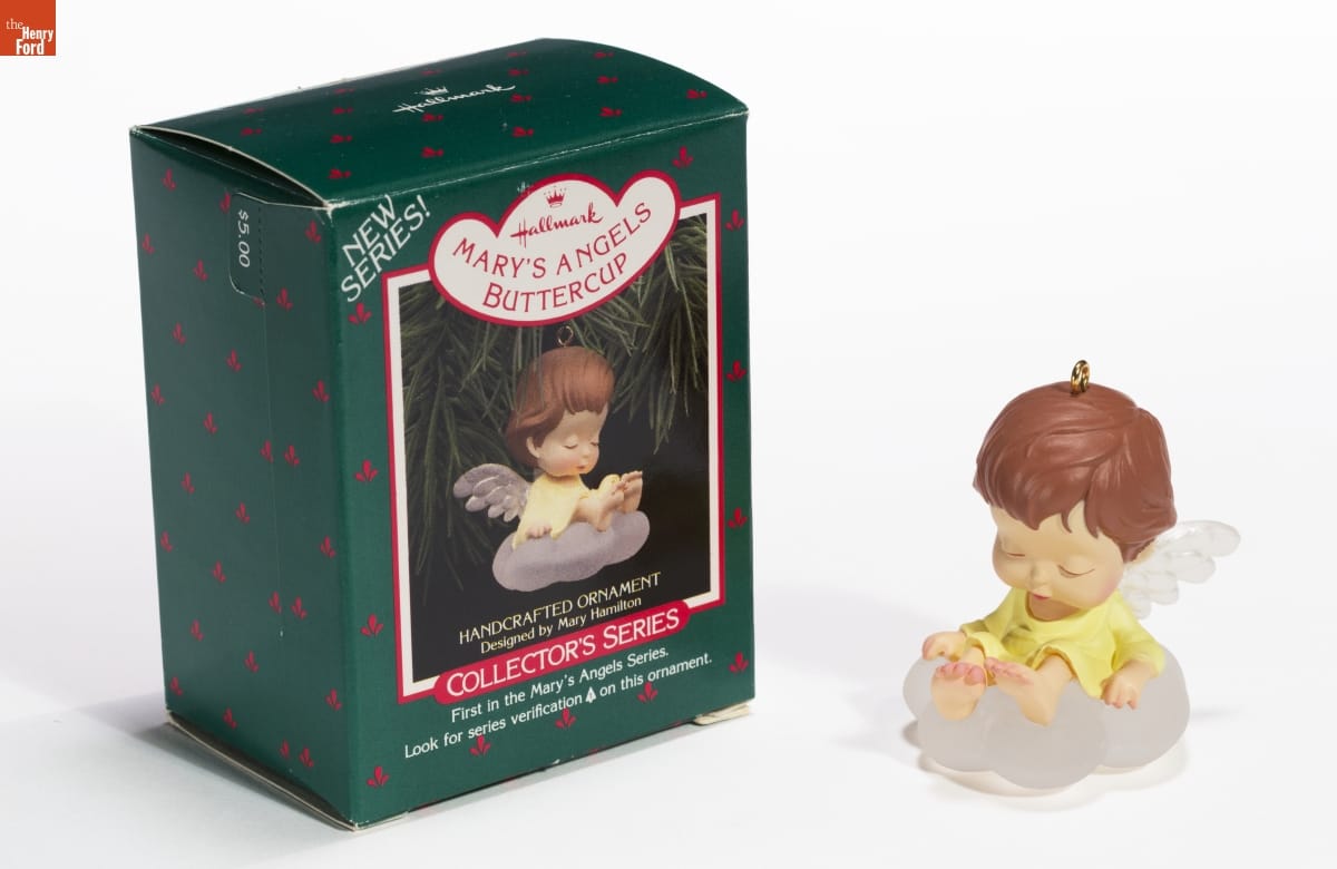 Ornament with large-headed , brown-haired child/angel wearing a yellow dress with transparent wings sitting on transparent cloud; green box with image of ornament sits next to it