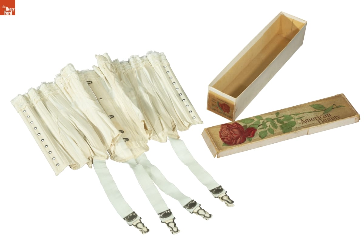 White corset with hook-and-eyes and belts with clips hanging down, next to box with image of red rose and text 