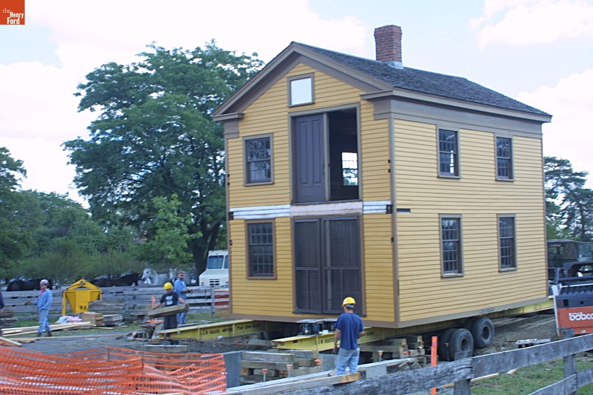 Richart Wagon Shop Being Relocated during the Greenfield Village Restoration Project, September 2002 Two-story yellow wooden building on rails and wheels on a construction site with people in hard hats nearby