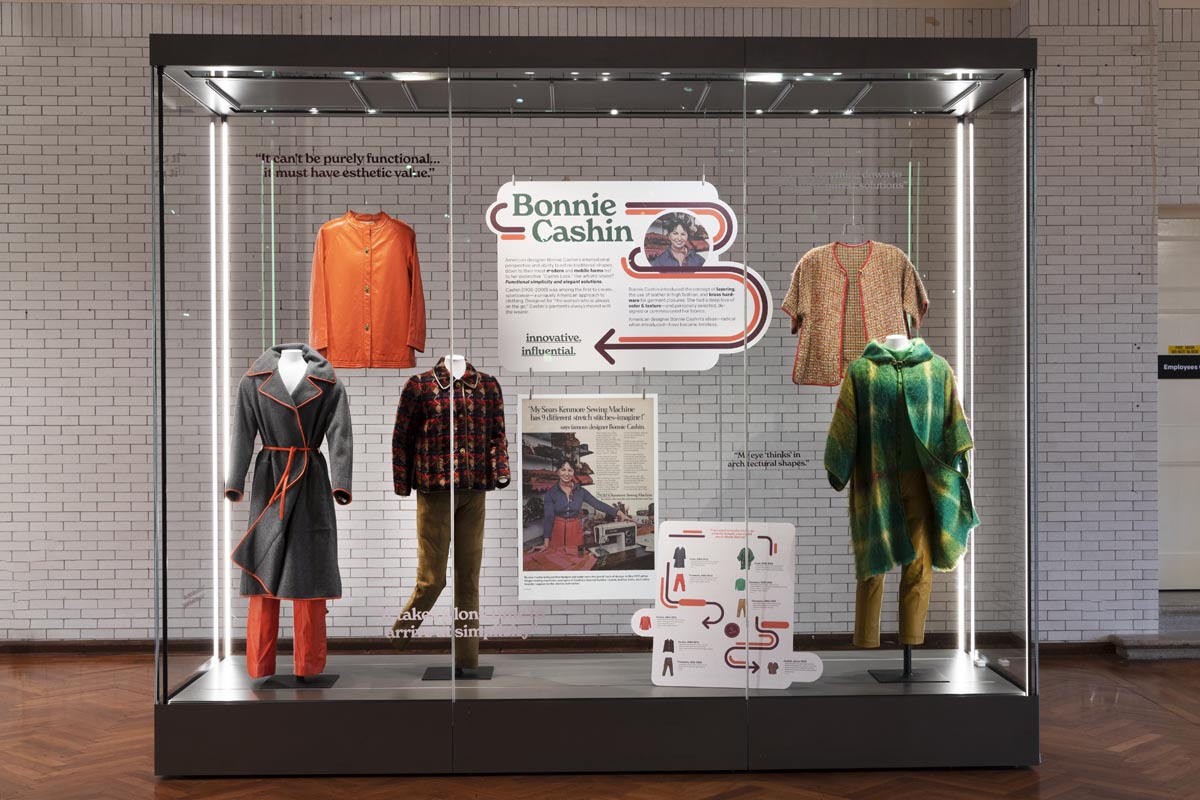 Large glass display case containing mannequins wearing clothing, signs and graphics, and additional hung clothing