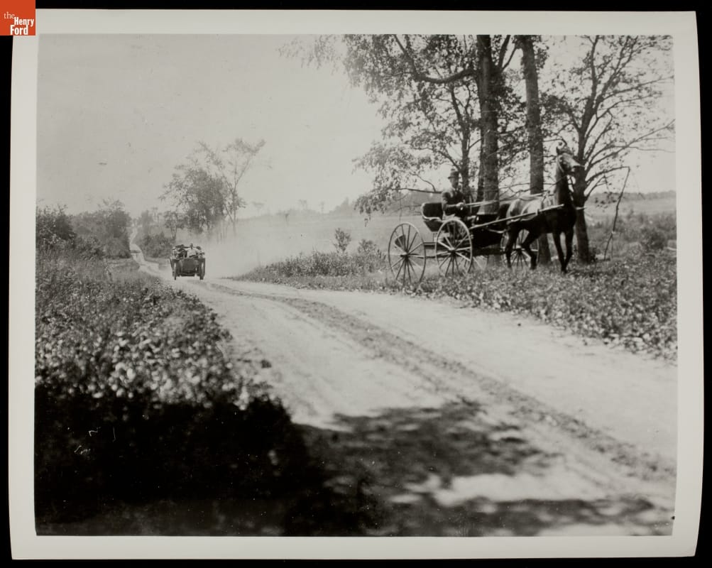 Car in distance on dirt road between fields; a horse-drawn carriage is pulled over on one side