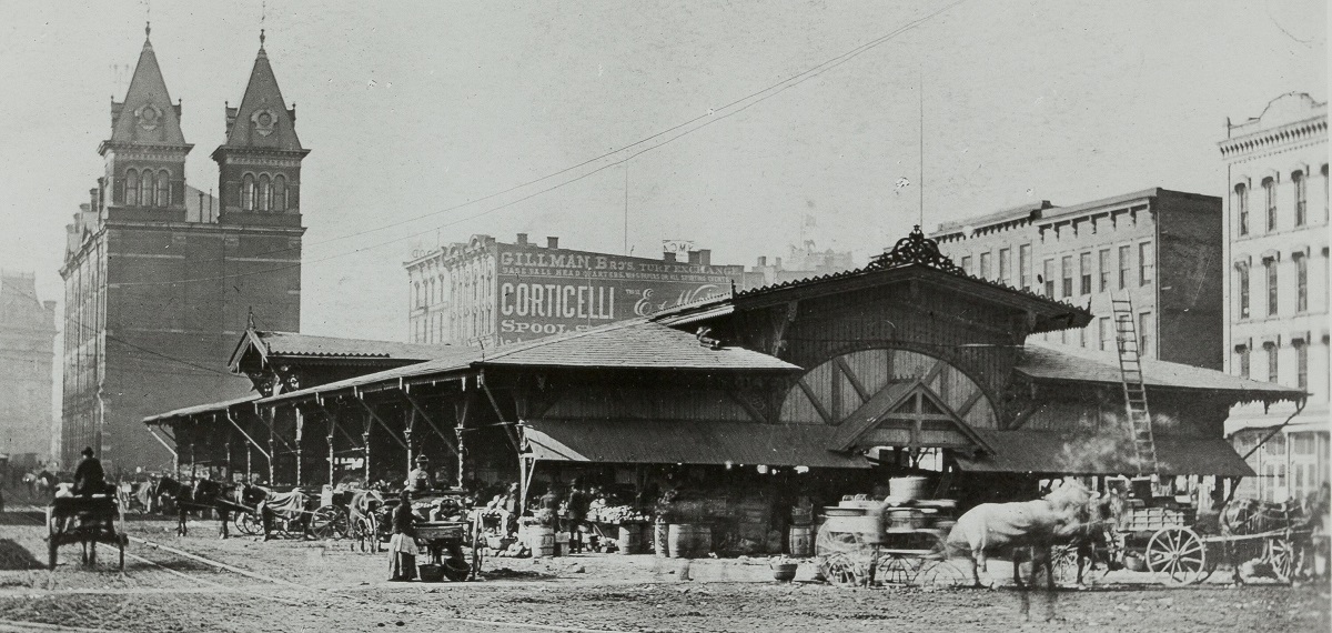 Black-and-white photo of long, low, open building with people and horses and carriages around it