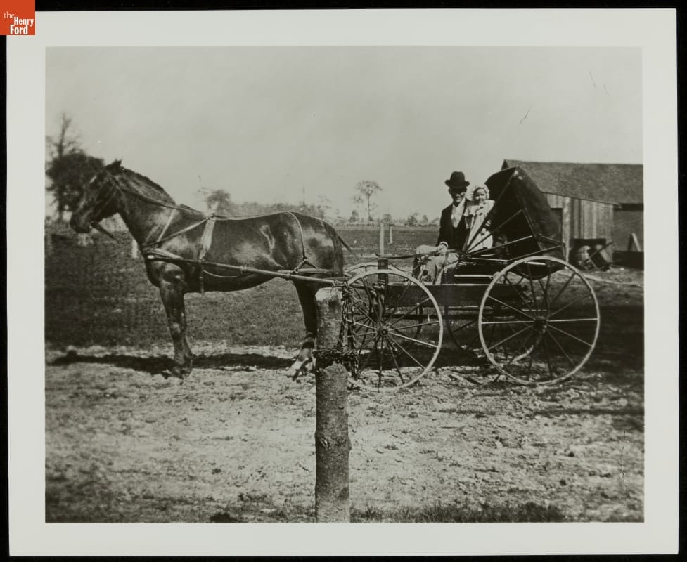 Man in suit and hat with child in an open buggy hitched to a horse, in front of a field 