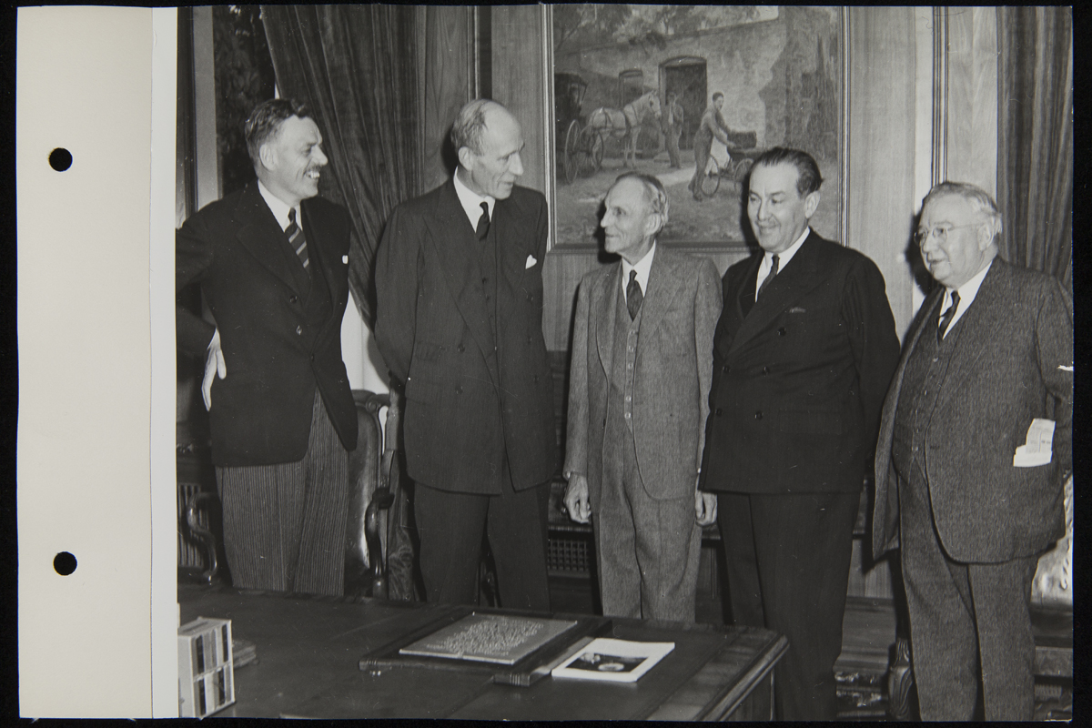 Five men in suits stand behind a desk in an office