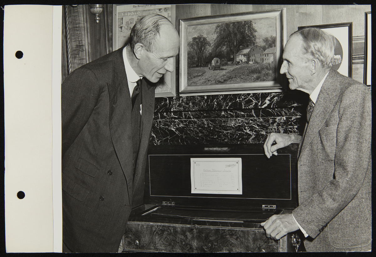 Two men talk over an open box in an office; one peers in