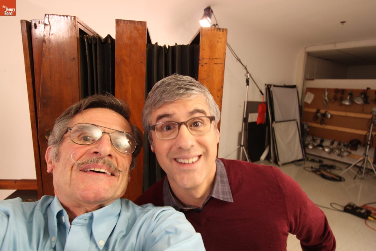 Two men smile smile for a selfie with a large old-fashioned box camera and modern photography equipment behind them