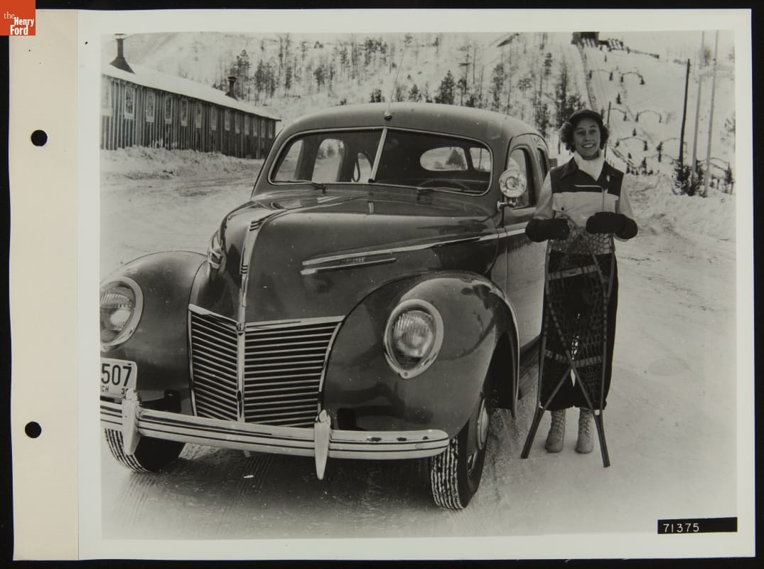 Woman, holding two snowshoes, stands next to a car with a snowy hill (perhaps a ski slope) in the background and a long low wooden building to one side