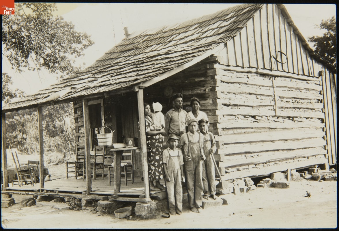 Indian Relief Project, McCurtain, Oklahoma, June 18, 1934 Black-and-white photo of group of adults and children standing on or near porch of very minimalistic wooden house