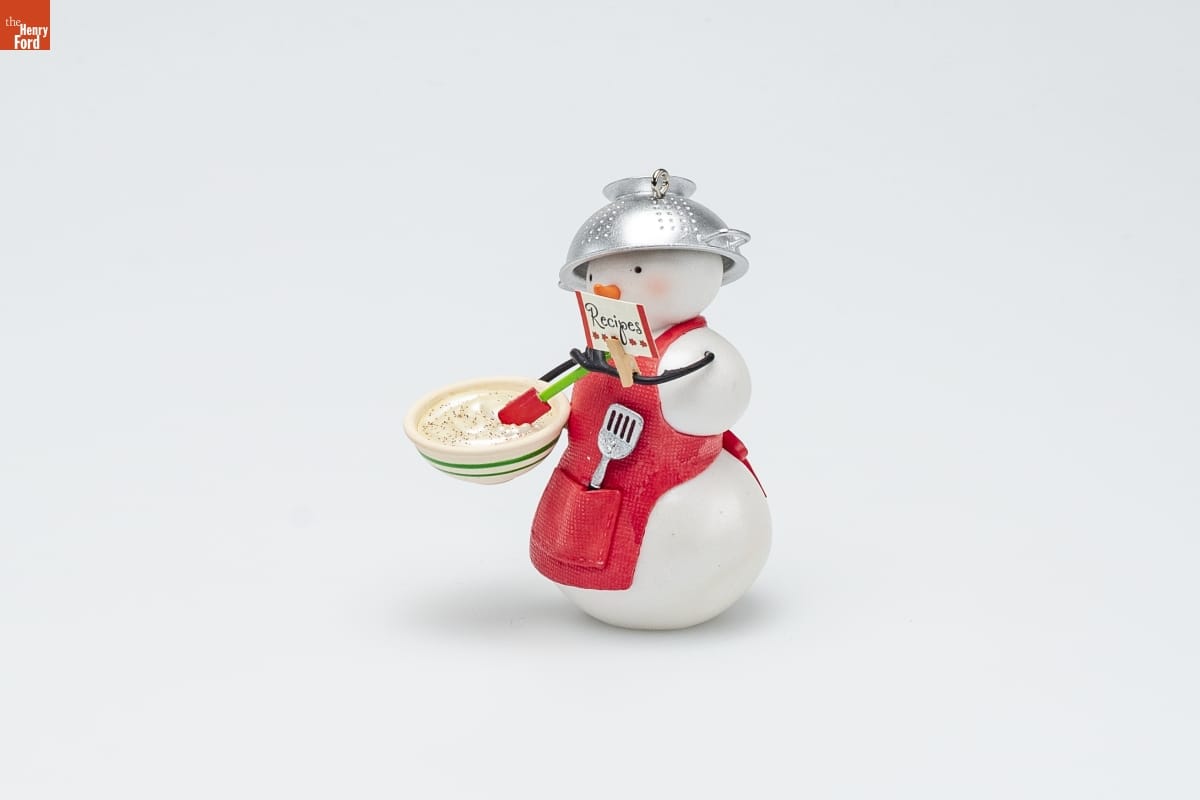 Ornament of a snowman wearing a red apron with a spatula in the pocket and a colander on its head, holding a bowl of dough in one hand and a card labeled 