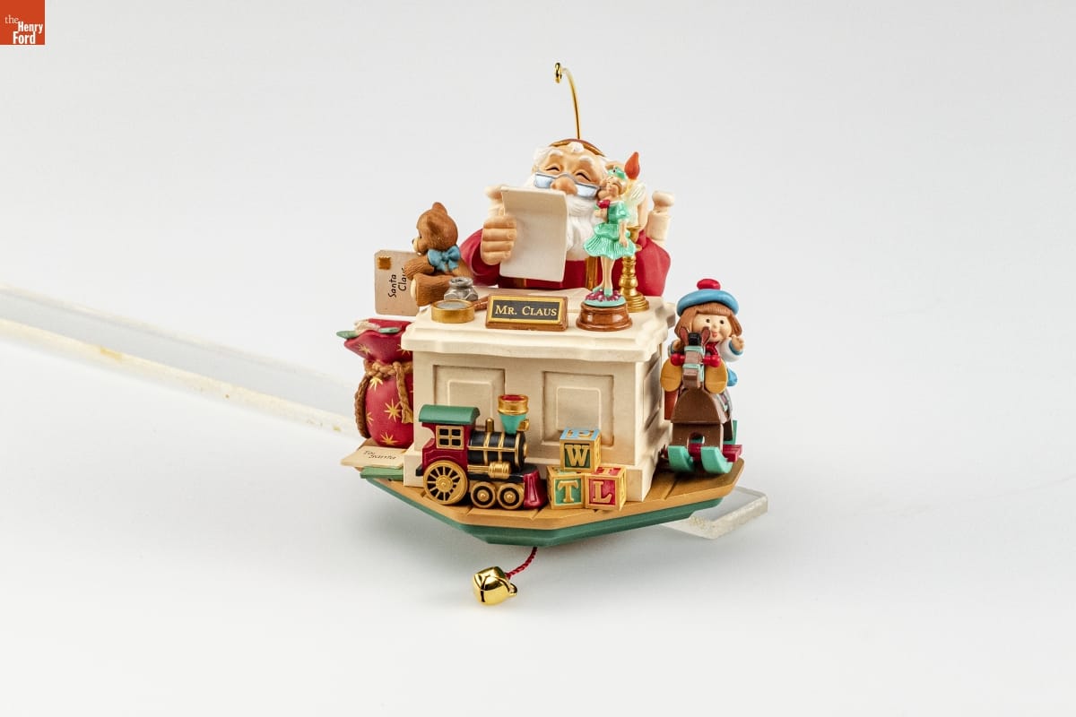Ornament depicting Santa Claus sitting behind a desk with sign 