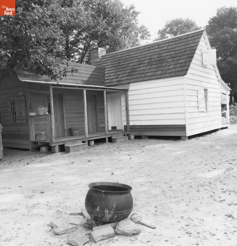 Black-and-white photo of wooden house with large iron kettle over ring of stones in dirt yard