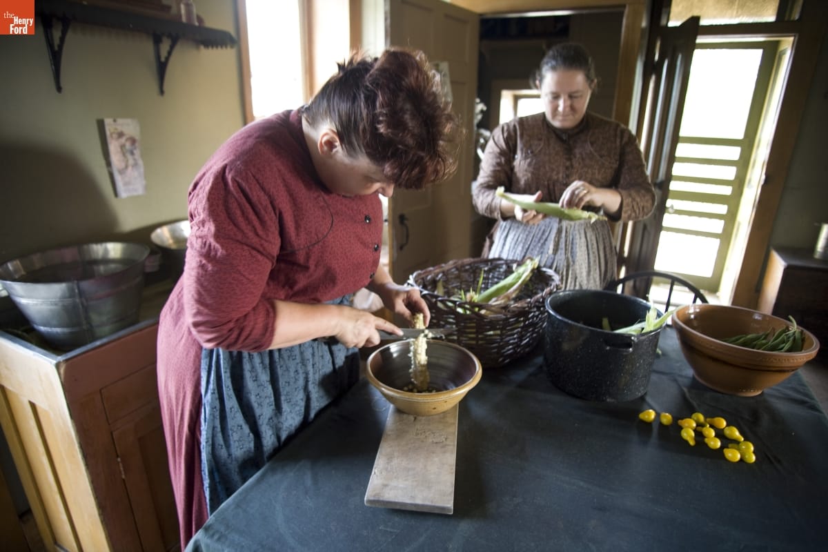 Two women shuck and remove kernels from corn cobs at a kitchen table containing other dishes and food