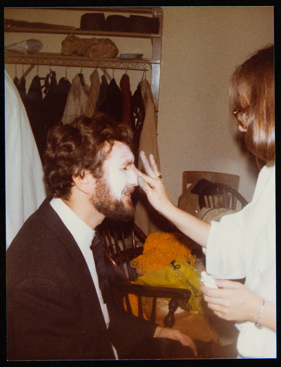 Seated man having makeup applied by a standing woman