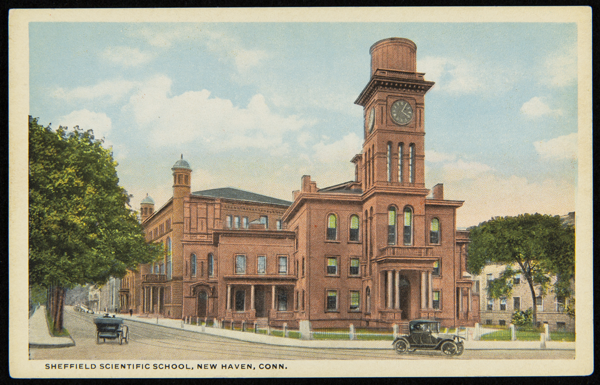 Postcard showing red brick building with cars passing it on street; also contains text