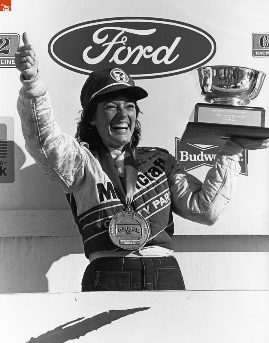 Lyn St. James at IMSA, Watkins Glen, NY, 1985 Smiling woman in a jumpsuit and baseball cap, with large medal around her neck, holds a trophy in one hand and makes a thumbs-up with the other, in front of a wall with Ford and Budweiser logos, among others