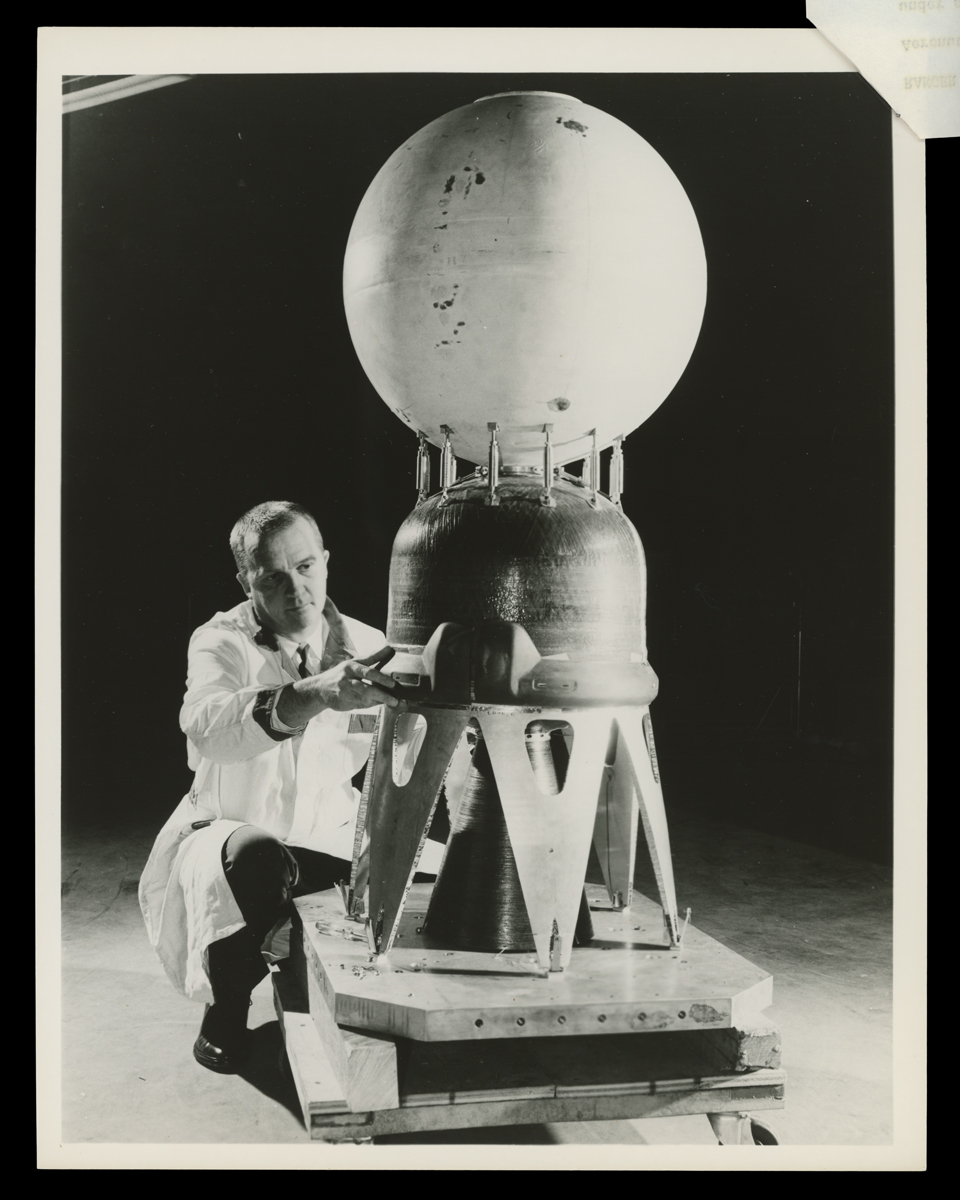 Man in lab coat kneels, working on a piece of equipment with stands, a dome-shaped middle portion, and a sphere at the top