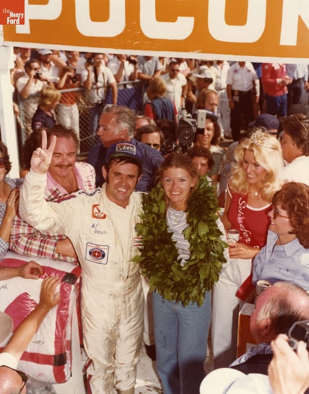 Man in white jumpsuit with arm around woman with large laurel wreath around her neck stand surrounded by other people, folded quilt to one side