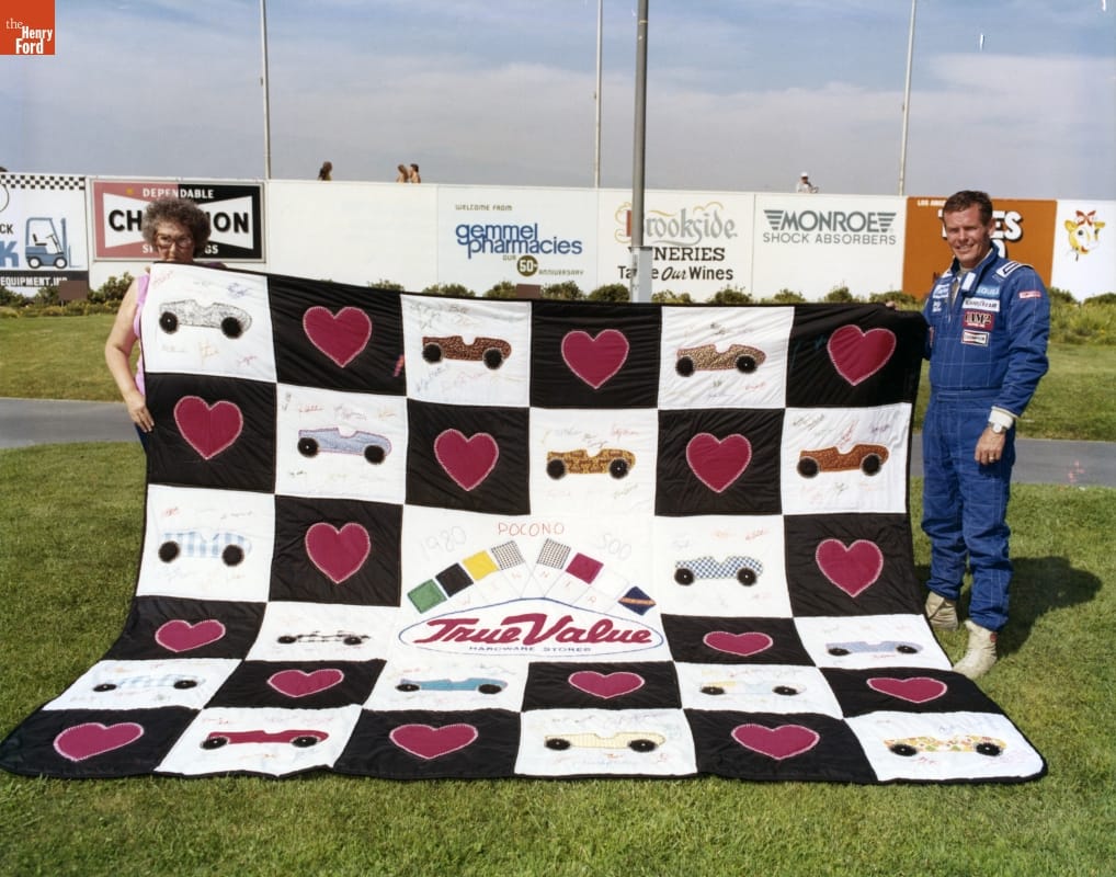 Man in blue jumpsuit and woman with curly hair wearing tortoiseshell glasses hold corners of a quilt spread out on the grass