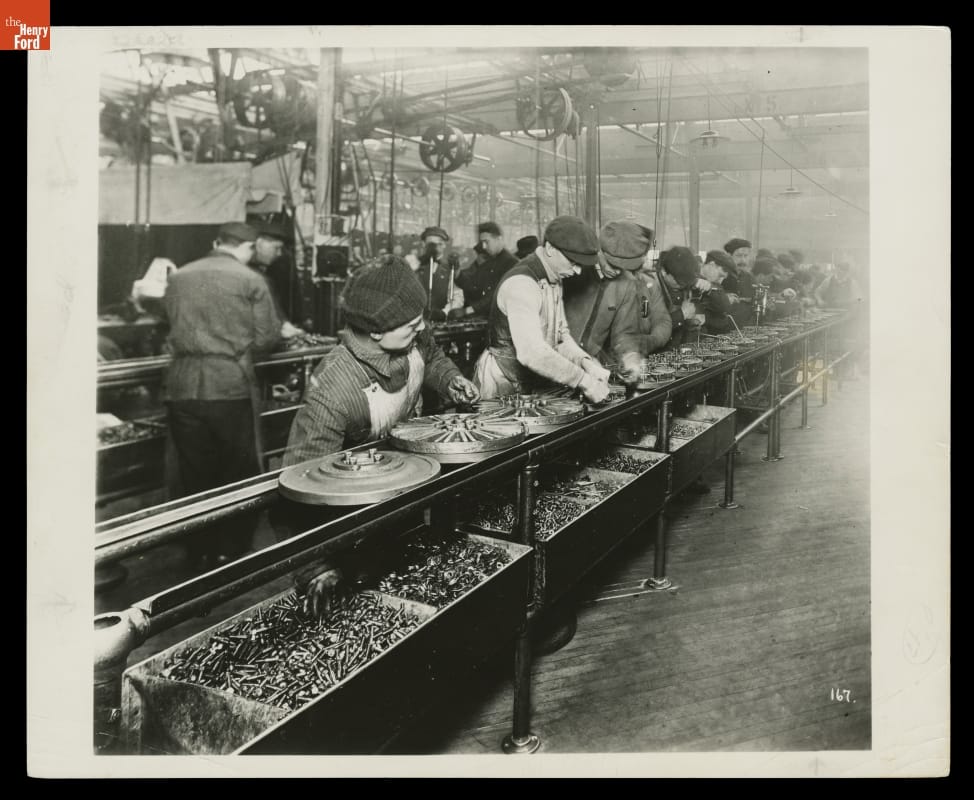 People work in a factory at a long line containing round parts, with bins of screws or bolts underneath