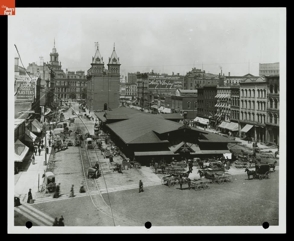 City scene with large, low building in center of photo with people and horse carriages nearby