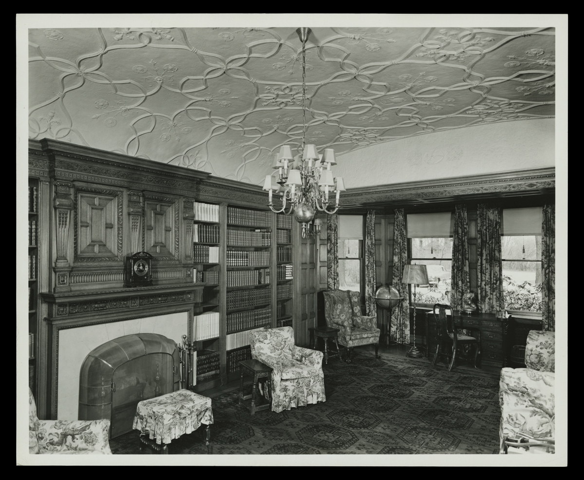 Room interior with carved wood, ornately patterned ceiling, bookcases, and upholstered chairs among other furniture