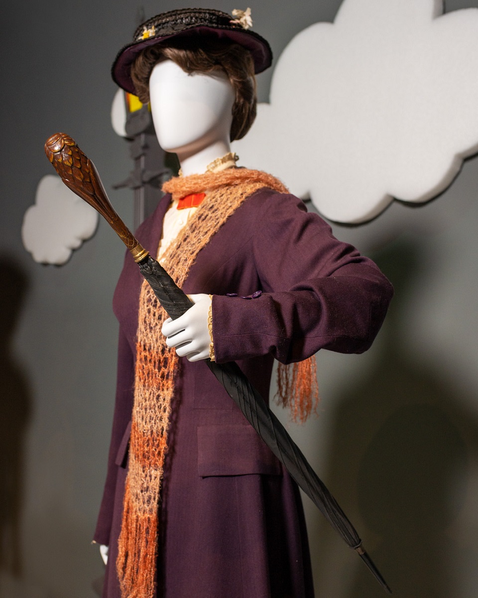 Costume from Mary Poppins in the Heroes and Villains: The Art of the Disney Costume exhibit Mannequin dressed in a dark purple coat with an orange knitted scarf, black hat, and umbrella with carved wooden handle, posed in front of a gray wall with white clouds